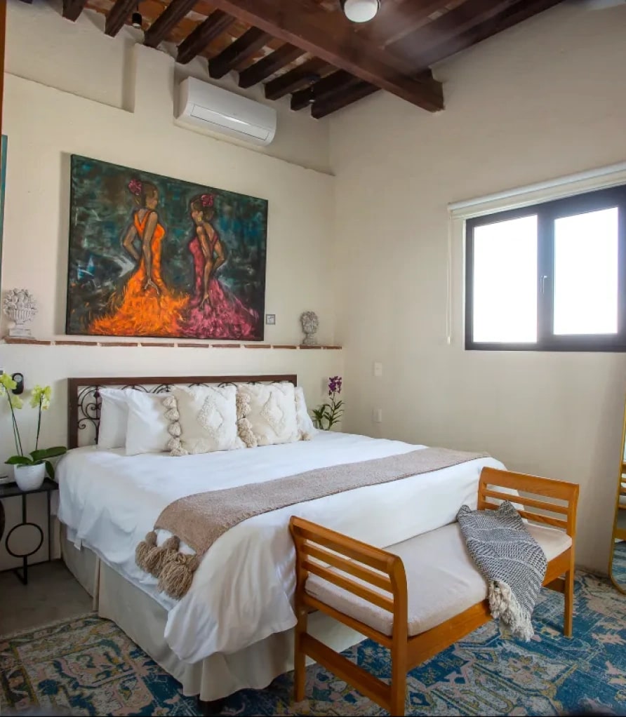 King bed with a wrought-iron headboard and a timber bench at the foot, under beams, with a painting of two dancers on the wall