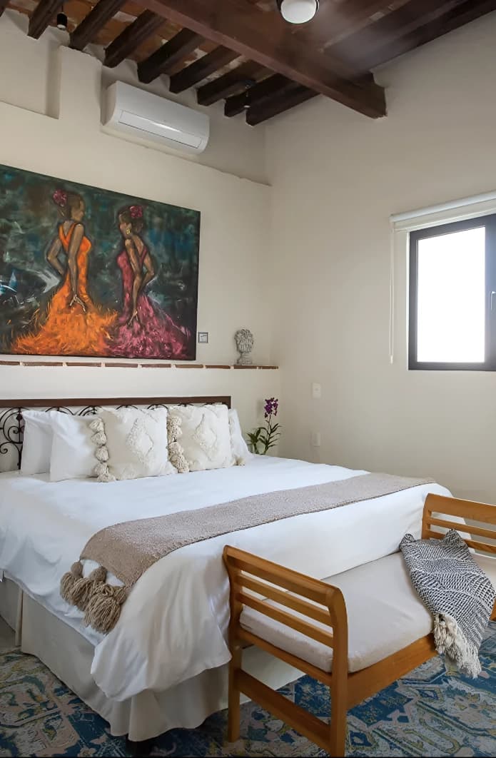 King bed with a wrought-iron headboard and a timber bench at the foot, under beams, with a painting of two dancers on the wall