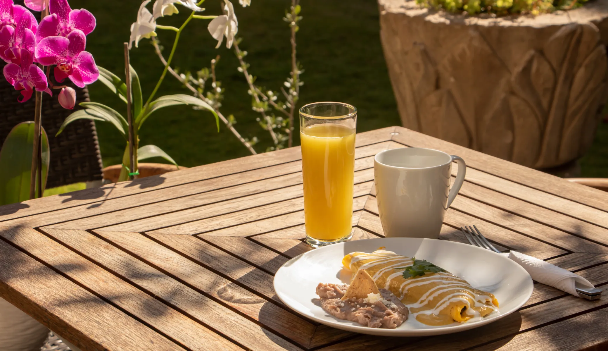 Breakfast served on a timber table in the garden: enchiladas in sauce, refried beans, orange juice and coffee, beside orchids