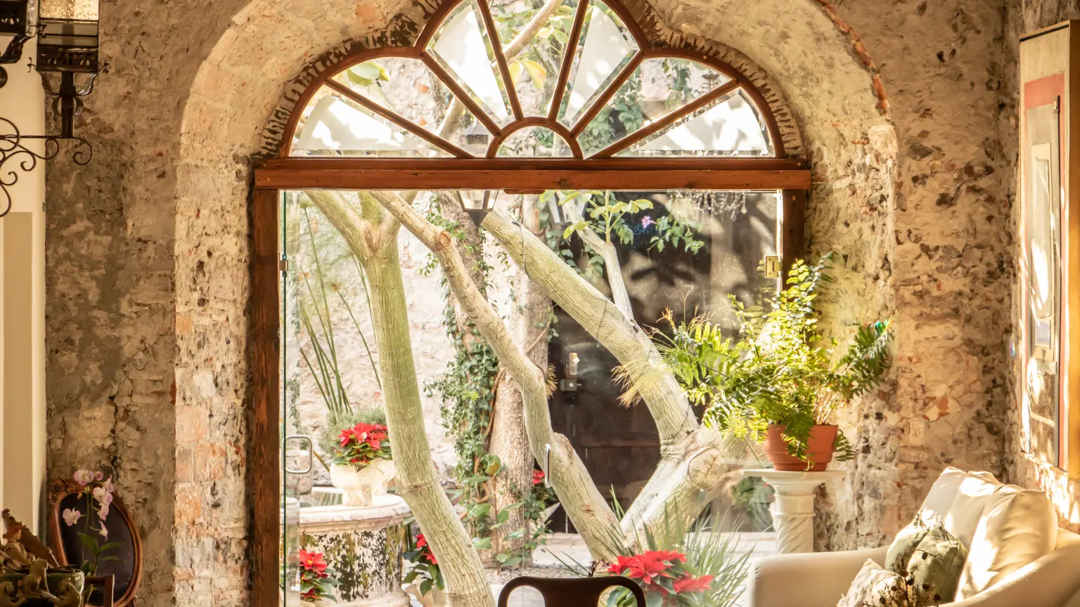 The living room seen through a stone arch with a timber fanlight, toward the courtyard with trees and poinsettias