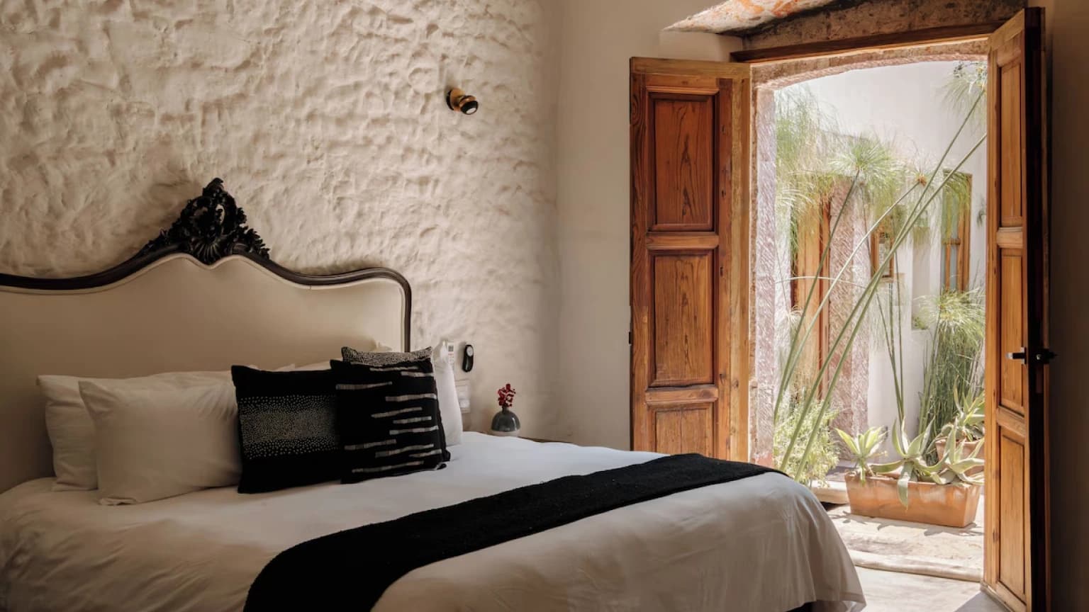 A bedroom with a carved headboard, timber doors open onto a courtyard with papyrus and agaves