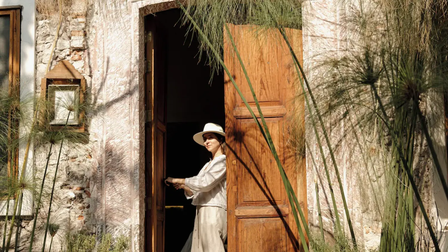 A guest in a hat steps out through the timber door, among the courtyard's papyrus