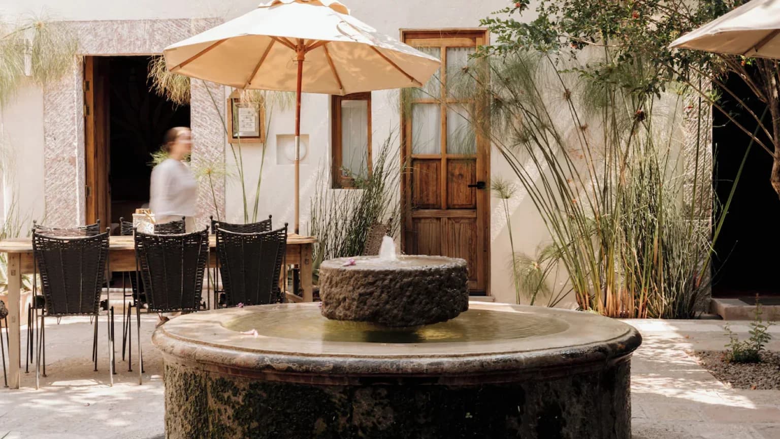 The courtyard's cantera fountain, with a guest passing by and the table under its umbrella beyond