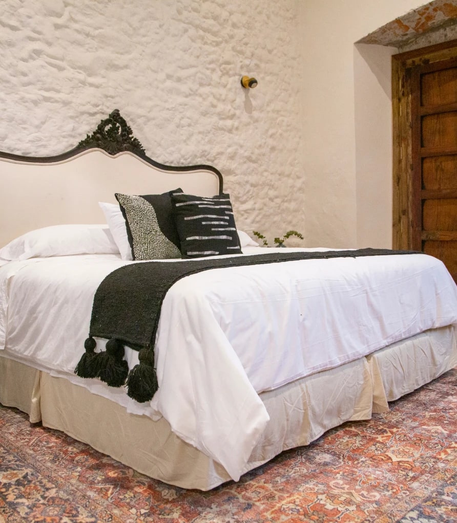 King bed with an upholstered headboard and a carved crest, a black runner with tassels and a whitewashed stone wall