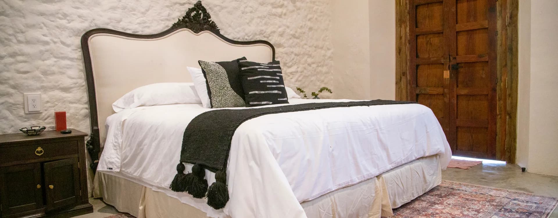 King bed with an upholstered headboard and a carved crest, a black runner with tassels and a whitewashed stone wall