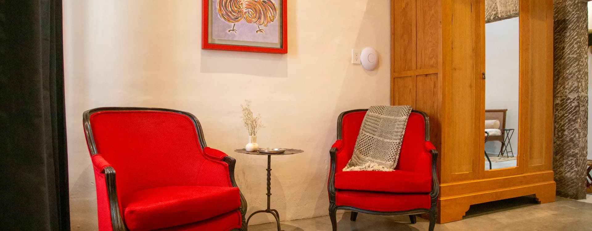 Two red armchairs and a side table beside a timber wardrobe and a framed print of a rooster
