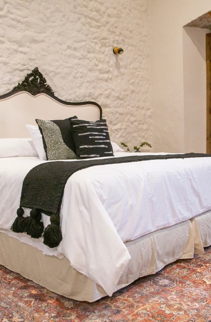 King bed with an upholstered headboard and a carved crest, a black runner with tassels and a whitewashed stone wall