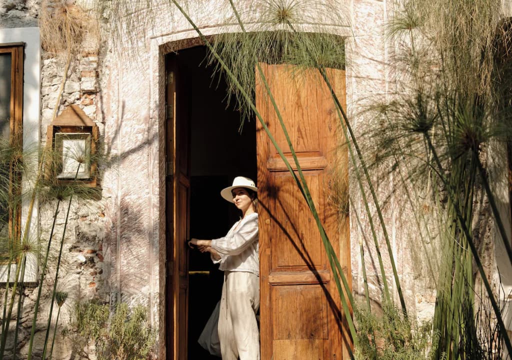 Someone in light linen and a brimmed hat in the wooden doorway, among papyrus and agaves