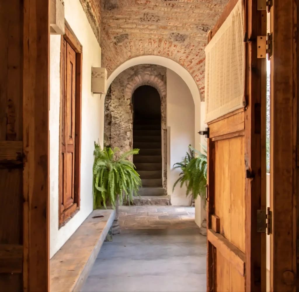 The vaulted brick corridor of the house, with timber doors, a stone bench and the stone staircase beyond, under an arch