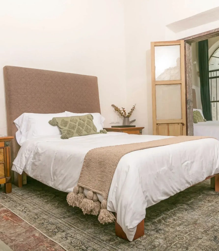 Bed with an upholstered headboard and a green cushion, a timber nightstand and a Persian rug, with a glass door beyond