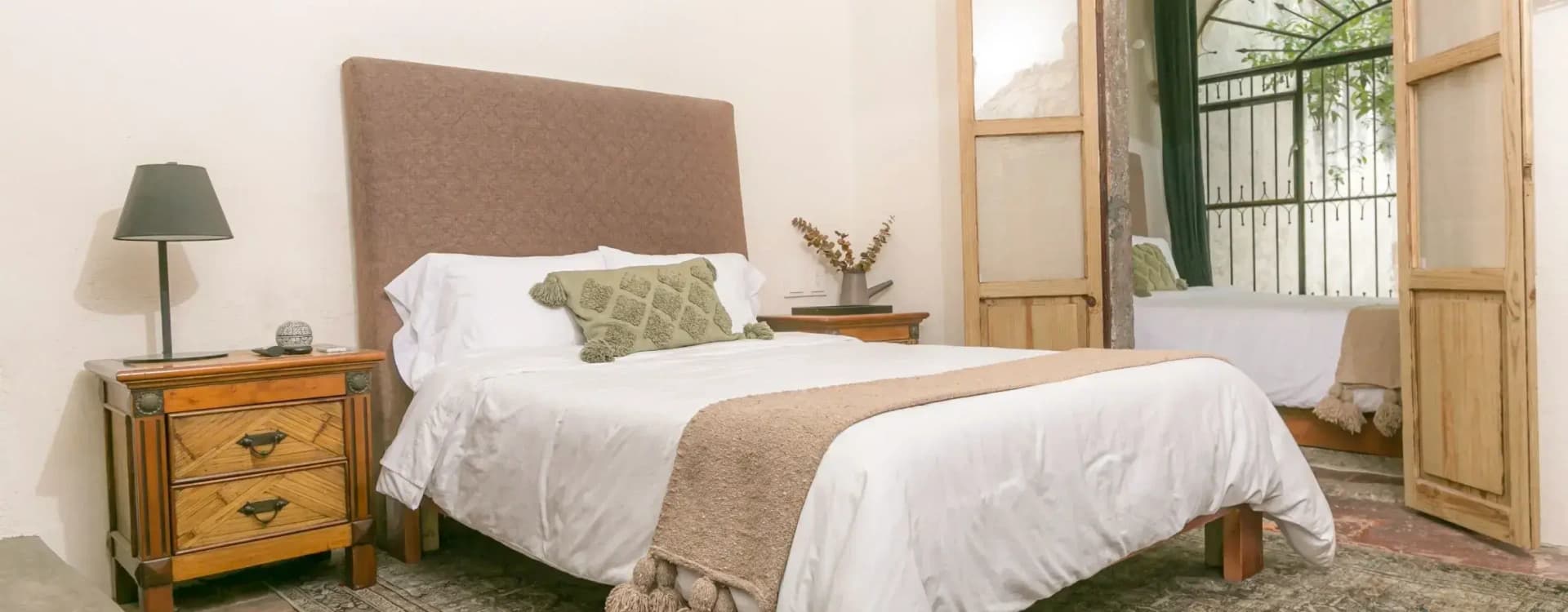 Bed with an upholstered headboard and a green cushion, a timber nightstand and a Persian rug, with a glass door beyond