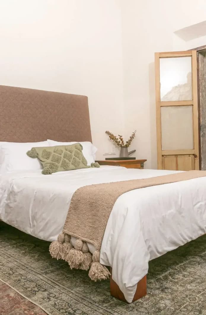 Bed with an upholstered headboard and a green cushion, a timber nightstand and a Persian rug, with a glass door beyond