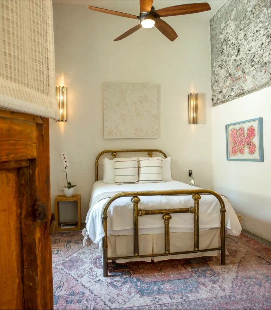 Room with a brass queen bed between two metal sconces, a Persian rug and a stone wall in the corner