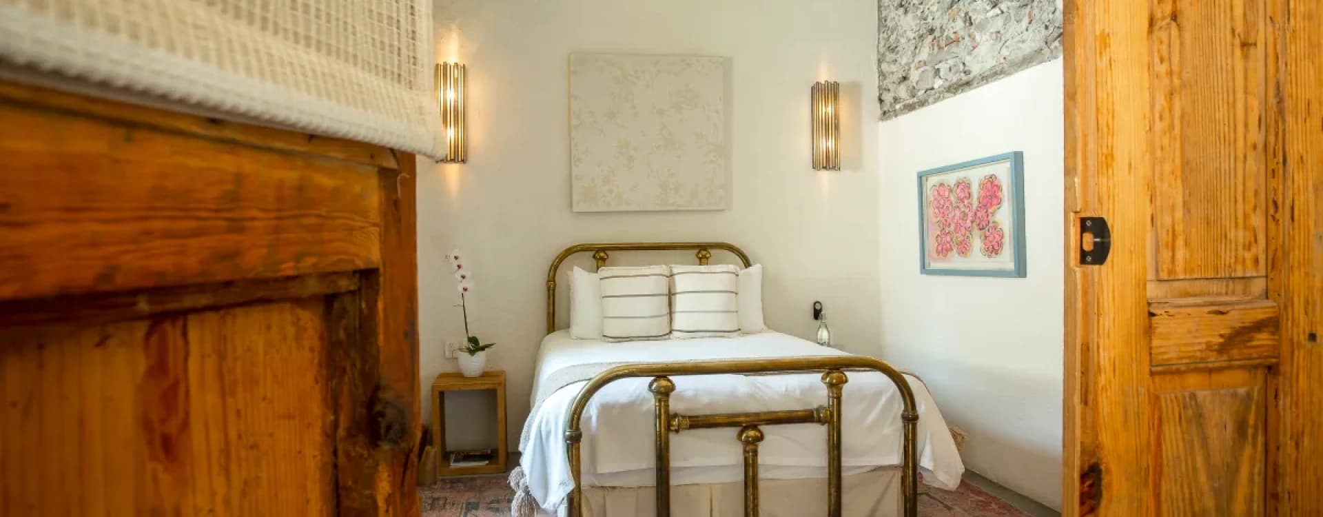 Room with a brass queen bed between two metal sconces, a Persian rug and a stone wall in the corner