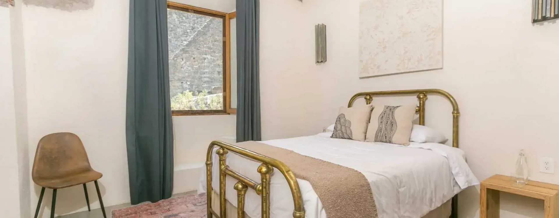 Brass queen bed with a wool runner with tassels, beside a window with blue-grey curtains