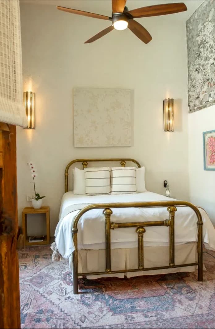 Room with a brass queen bed between two metal sconces, a Persian rug and a stone wall in the corner