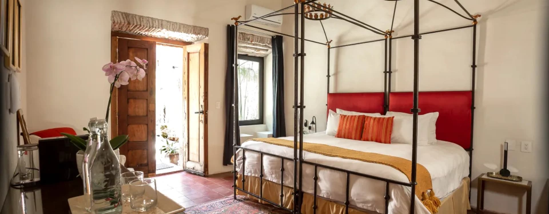 Suite with a wrought-iron four-poster king bed and a red headboard, under timber beams and beside the door to the patio