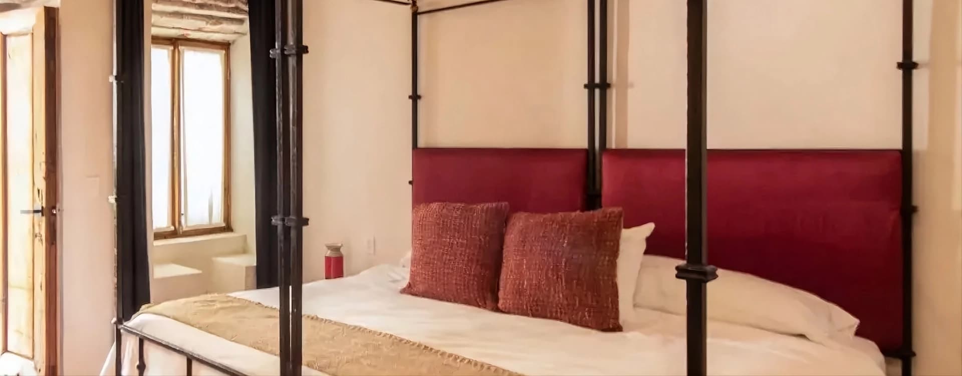 Wrought-iron four-poster king bed with a red headboard and a woven runner, beside a window