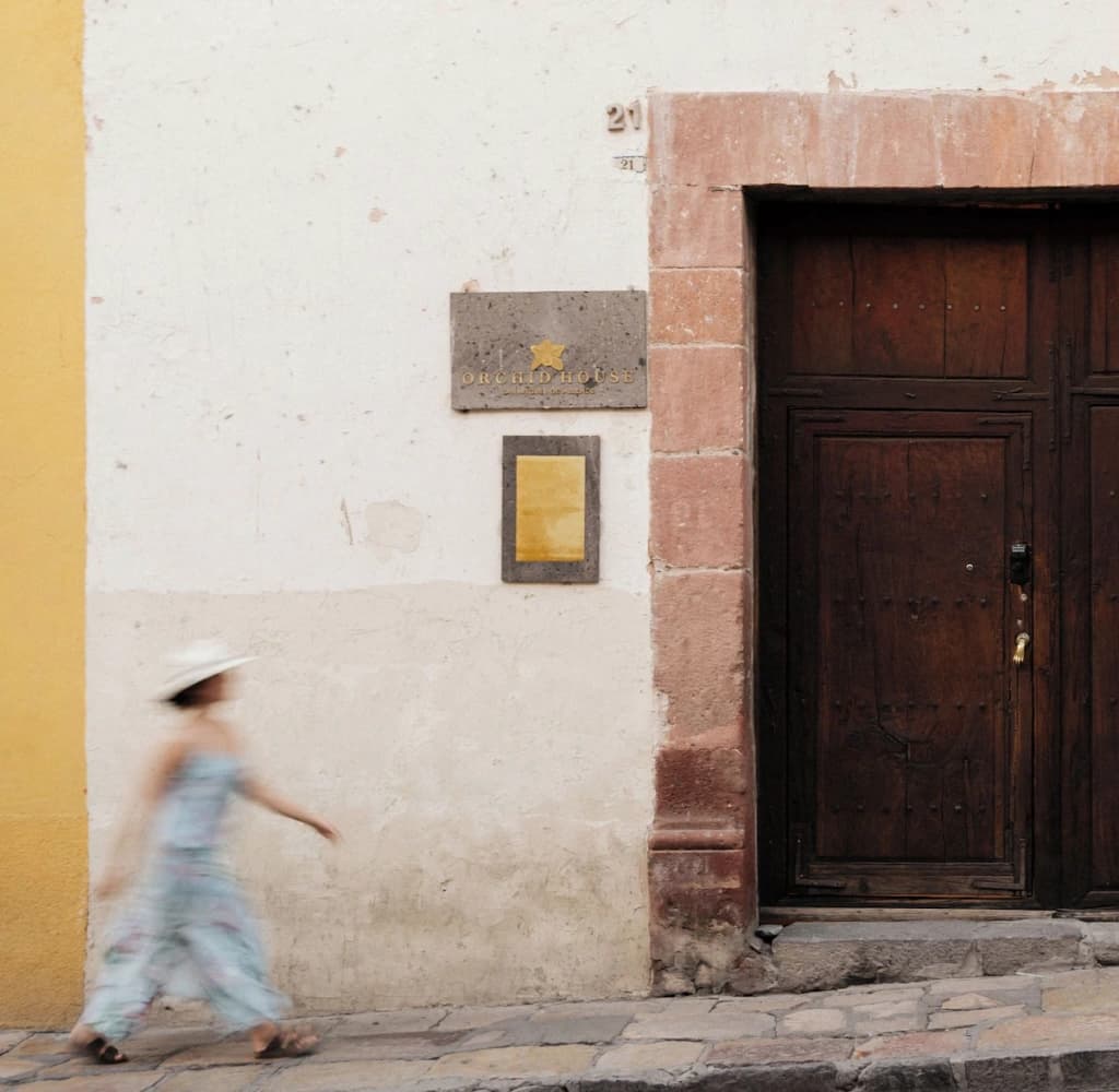 The house's facade on the street: the stone plaque with the gold orchid and the Orchid House name beside the wooden door, as a guest in a hat walks past